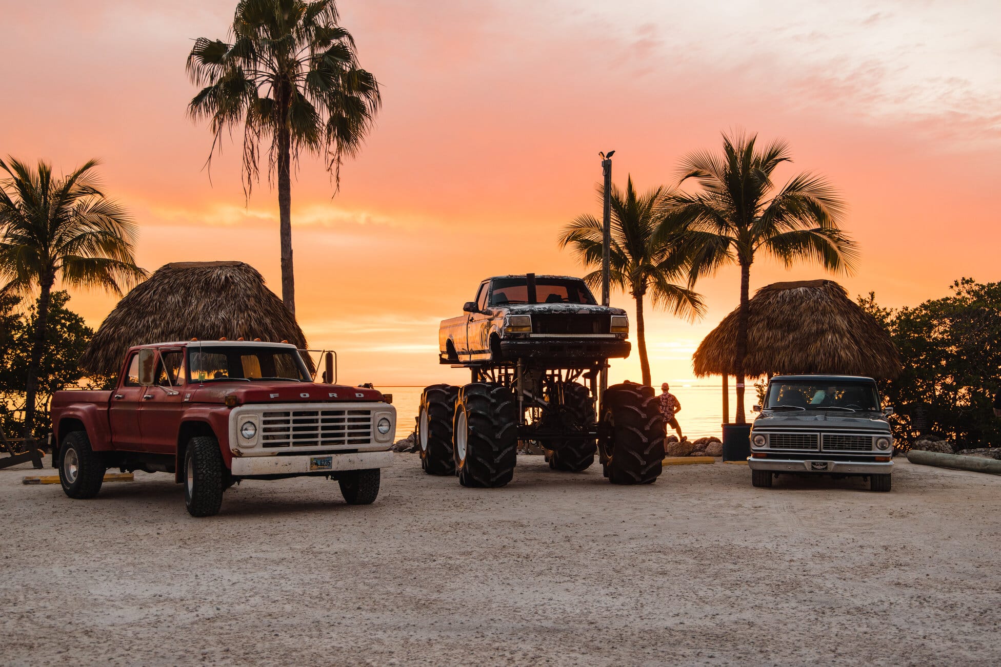 Trucks parked between palm trees and tiki huts under an orange sunset sky