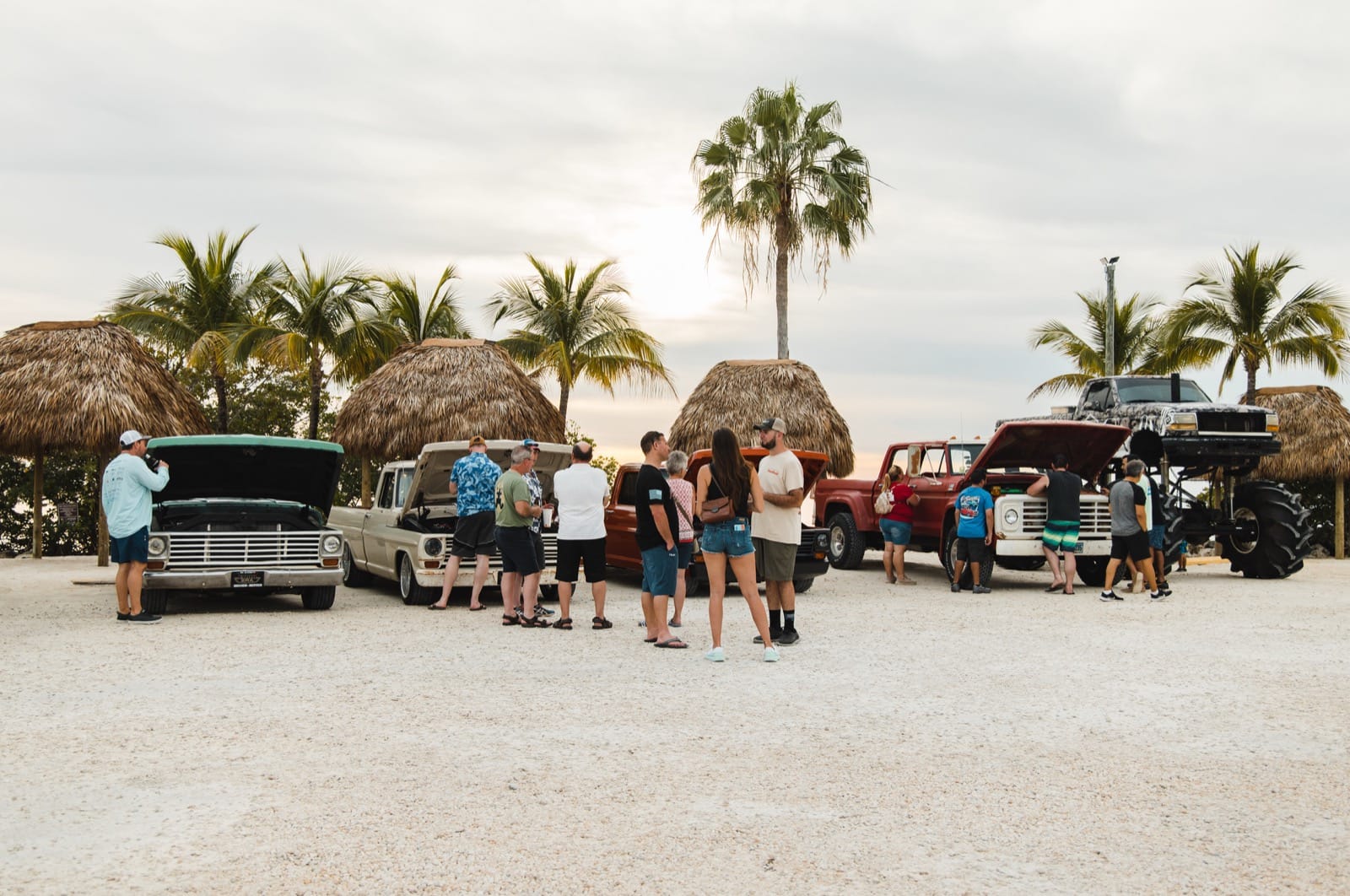 A crowd walking the sand between classic trucks, palms, and tiki huts